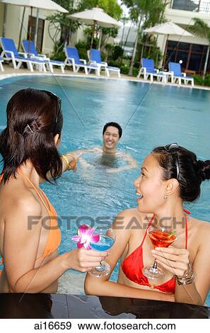 Young women in swimming pool, holding cocktails, looking at man swimming behind them View Large Photo Image Stock Photo - Young women in swimming pool, holding cocktails, looking at man swimming behind them . Fotosearch