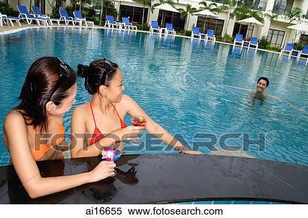 Young women in swimming pool, holding cocktails, turning to look at man swimming behind them View Large Photo Image Stock Photography - Young women in swimming pool, holding cocktails, turning to look at man swimming behind them . Fotosearch