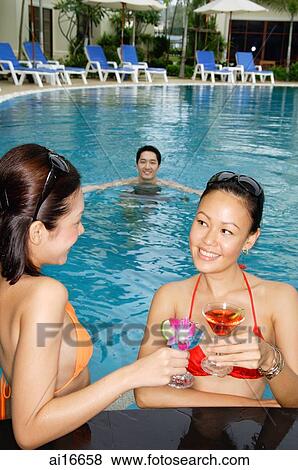 Young women talking in swimming pool, holding cocktails, man swimming behind them View Large Photo Image Stock Photo - Young women talking in swimming pool, holding cocktails, man swimming behind them . Fotosearch