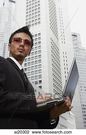 Stock Image - Businessman in sunglasses, standing with laptop, buildings behind him. Fotosearch