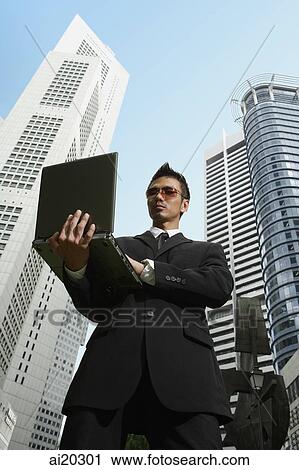 Businessman standing with laptop, buildings behind him View Large Photo Image Stock Image - Businessman standing with laptop, buildings behind him. Fotosearch