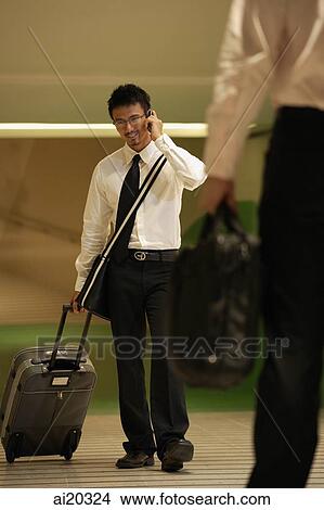 Businessman using mobile phone while pulling luggage behind him View Large Photo Image Picture - Businessman using mobile phone while pulling luggage behind him. Fotosearch