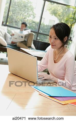 Stock Image - Couple at home, woman using laptop, man in background. Fotosearch