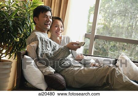 Couple in living room, sitting side by side on sofa, man holding TV remote control View Large Photo Image Stock Image - Couple in living room, sitting side by side on sofa, man holding TV remote control. Fotosearch