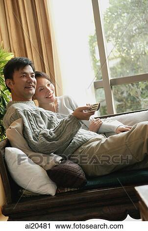 Couple sitting side by side on sofa, man holding TV remote control View Large Photo Image Stock Image - Couple sitting side by side on sofa, man holding TV remote control. Fotosearch