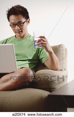 Man at home, using laptop, holding glass of water View Large Photo Image Picture - Man at home, using laptop, holding glass of water. Fotosearch