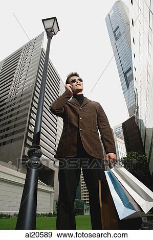 Man carrying shopping bags, using mobile phone, buildings in the background View Large Photo Image Stock Photo - Man carrying shopping bags, using mobile phone, buildings in the background. Fotosearch
