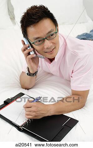 Man lying on bed, using mobile phone, notebook open in front of him View Large Photo Image Stock Image - Man lying on bed, using mobile phone, notebook open in front of him. Fotosearch
