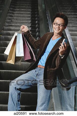 Man on escalator, carrying shopping bags and holding mobile phone, smiling at camera View Large Photo Image Stock Image - Man on escalator, carrying shopping bags and holding mobile phone, smiling at camera. Fotosearch