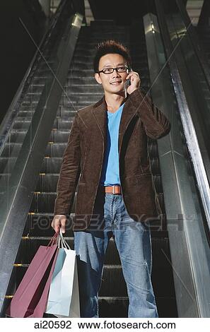 Stock Image - Man on escalator, carrying shopping bags, using mobile phone. Fotosearch