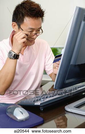 Man sitting in front of computer, looking at credit card View Large Photo Image Picture - Man sitting in front of computer, looking at credit card. Fotosearch