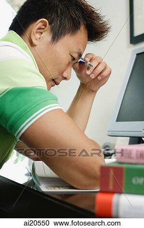 Stock Image - Man sitting in front of computer, holding a pen, looking down. Fotosearch