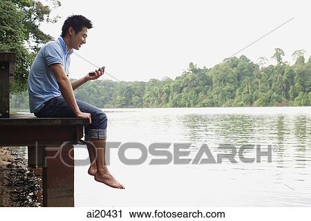 Stock Image - Man sitting on jetty, looking at mobile phone. Fotosearch
