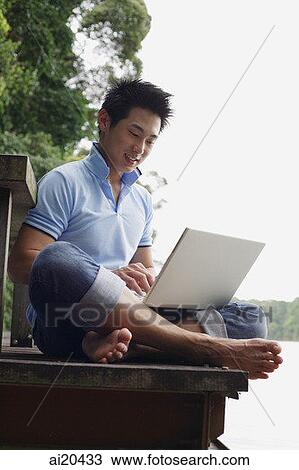 Stock Image - Man sitting on jetty, using laptop. Fotosearch