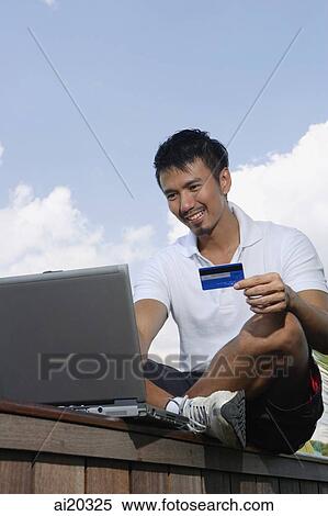 Man sitting outdoors, holding credit card and looking at laptop View Large Photo Image Stock Photography - Man sitting outdoors, holding credit card and looking at laptop. Fotosearch