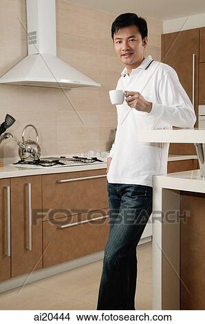 Man standing in kitchen, holding cup, looking at camera View Large Photo Image Picture - Man standing in kitchen, holding cup, looking at camera. Fotosearch