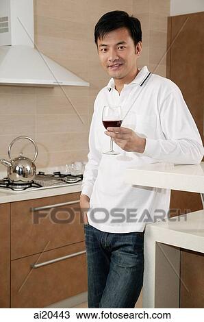 Stock Image - Man standing in kitchen, holding wine glass, looking at camera. Fotosearch