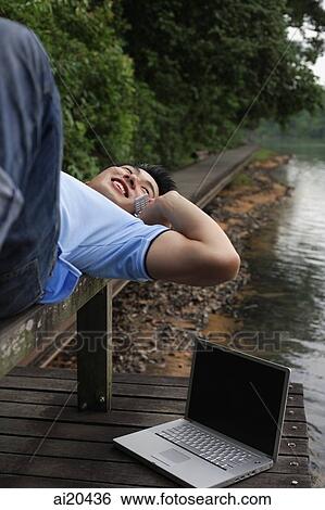 Man using mobile phone, lying on jetty, laptop next to him View Large Photo Image Stock Photograph - Man using mobile phone, lying on jetty, laptop next to him. Fotosearch