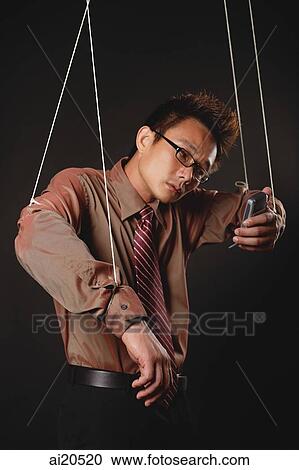 Stock Image - Man with marionette strings attached to his hands, holding mobile phone. Fotosearch