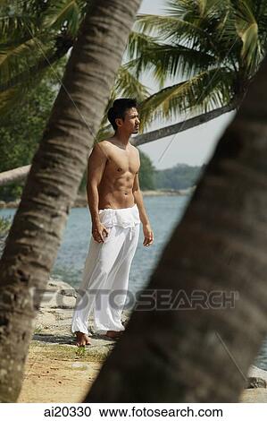 Stock Image - Shirtless man standing on the beach, looking away. Fotosearch