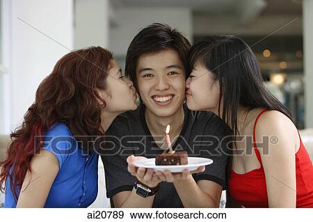 Stock Photo - Teenage boy in the middle holding plate with a cake and candle, girls on either side, kissing him on cheek. Fotosearch