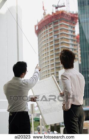 Stock Image - Two men looking at blueprint, one pointing to building in the background. Fotosearch