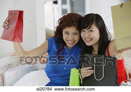 Two teenage girls sitting side by side holding up shopping bags View Large Photo Image Picture - Two teenage girls sitting side by side holding up shopping bags. Fotosearch