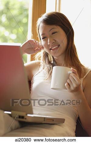 Stock Image - Woman holding mug, looking at laptop. Fotosearch