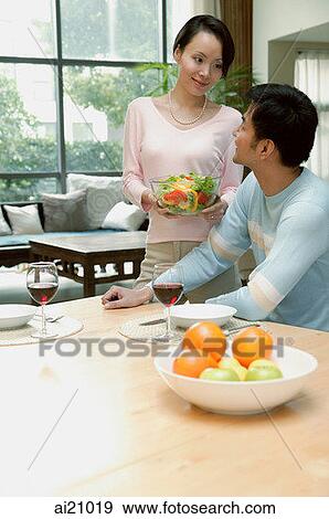 Stock Photo - Couple at home, man sitting at dining table, woman standing next to him holding bowl of salad. Fotosearch