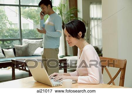 Couple at home, woman using laptop, man in the background, on the phone View Large Photo Image Stock Photo - Couple at home, woman using laptop, man in the background, on the phone. Fotosearch