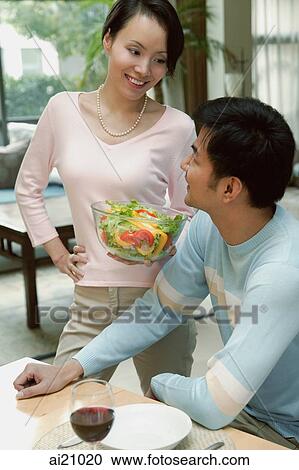 Man sitting at dining table looking at woman standing next to him holding bowl of salad View Large Photo Image Stock Image - Man sitting at dining table looking at woman standing next to him holding bowl of salad. Fotosearch
