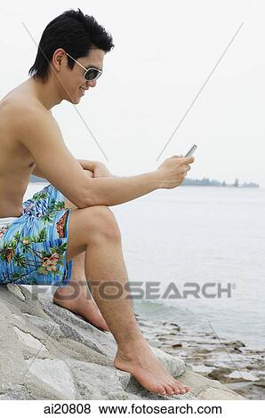 Man sitting by beach, looking at mobile phone View Large Photo Image Stock Photo - Man sitting by beach, looking at mobile phone. Fotosearch