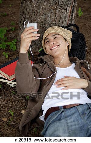 Young man lying at base of tree, listening to MP3 player View Large Photo Image Stock Photo - Young man lying at base of tree, listening to MP3 player. Fotosearch