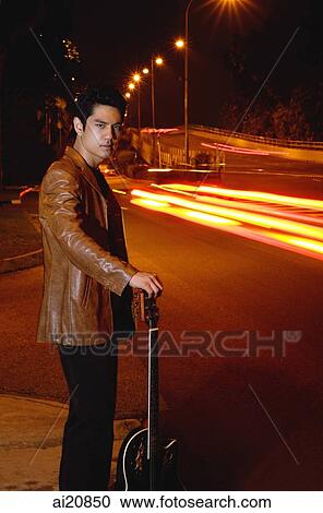 Stock Image - Young man wearing leather jacket, standing by road, leaning on guitar. Fotosearch