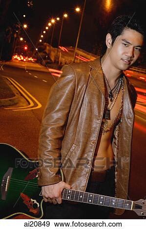 Stock Photo - Young man wearing leather jacket, standing by road, carrying guitar. Fotosearch