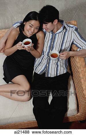 Stock Photo - Couple sitting on sofa, holding cups of tea. Fotosearch