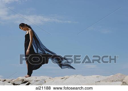 Stock Photo - Woman in black dress and scarf, standing on rocks, looking down. Fotosearch