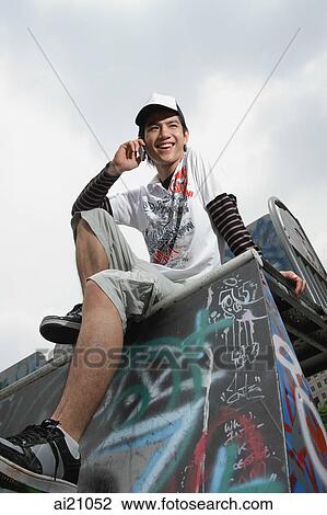 Stock Image - Young man using mobile phone, sitting on graffiti covered ledge. Fotosearch
