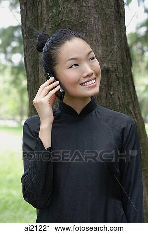 Stock Image - Young woman in traditional Chinese costume, standing next to tree, using mobile phone. Fotosearch