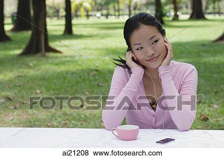 Young woman sitting at outdoor table, listening to MP3 player, smiling View Large Photo Image Stock Photo - Young woman sitting at outdoor table, listening to MP3 player, smiling. Fotosearch