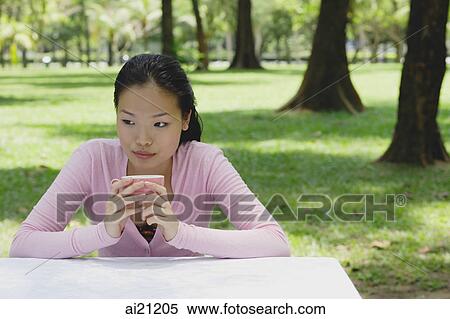 Stock Photography - Young woman sitting at outdoor table, holding coffee cup, looking away. Fotosearch