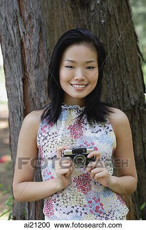 Young woman standing next to tree trunk, holding a camera, smiling View Large Photo Image Stock Image - Young woman standing next to tree trunk, holding a camera, smiling. Fotosearch