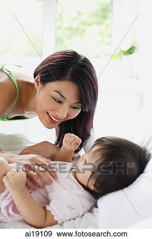 Stock Photo - Baby girl lying on bed, mother leaning over her. Fotosearch