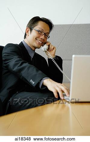 Stock Photo - Businessman in office using phone, looking at laptop. Fotosearch