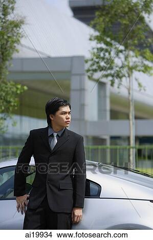 Picture - Businessman leaning on silver sports car, looking away. Fotosearch
