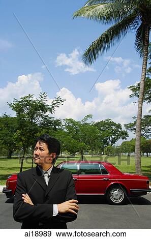 Stock Photo - Businessman standing with arms crossed, looking away, red car in the background. Fotosearch