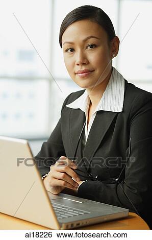Stock Photograph - Businesswoman sitting in front of laptop, looking at camera, portrait. Fotosearch