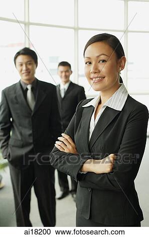 Stock Image - Businesswoman with arms crossed, two businessman in the background. Fotosearch