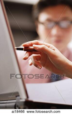 Stock Photograph - Close up of woman's hand, holding pen, pointing at laptop, man in the background. Fotosearch