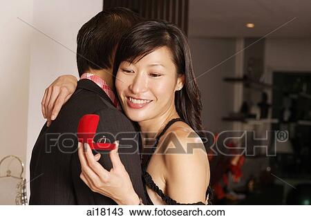 Stock Image - Couple embracing, woman looking at ring in box. Fotosearch